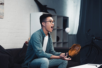 Wall Mural - emotional young asian man with baseball glove and ball sitting on couch and watching sport match at home