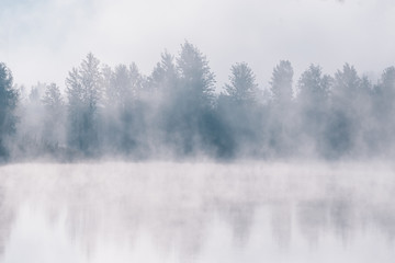 Foggy river coast at the autumn morning.  Reflections of forest trees in the water.