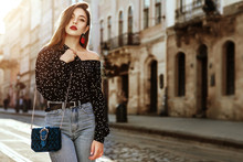 Outdoor Portrait Of Yong Beautiful Fashionable Woman Wearing Stylish Black White Polka Dot Blouse, Blue Jeans, Red Earrings, With Small Quilted Bag. Model Posing In Street Of European City. Copy Space