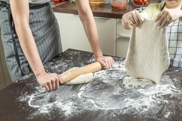 Wall Mural - Two adult girl and the child cook dough. Dough is spread out on the big table sprinkled with flour. The child has lifted the rolled dough over a table, and the girl rolls dough with a rolling pin