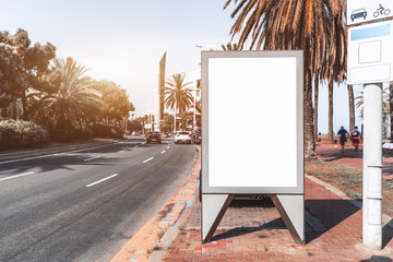 Outdoor empty informational board placeholder; white blank city billboard mock-up near the road; vertical blank advertising banner template on the sidewalk between the embankment and the highway