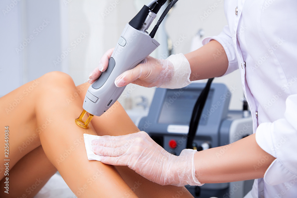Close-up of a female cosmetologist in a medical coat making a young woman  a procedure laser hair removal for leg. Cosmetology, ionization, diamond procedures. - obrazy, fototapety, plakaty 