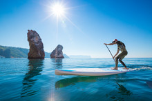 Young Man Having Fun Stand Up Paddling In The Sea. SUP. Guy Training In The Morning On Paddle Board Near The Rocks.