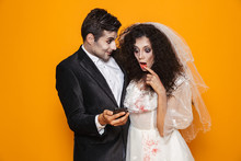 Photo Of Excited Zombie Couple Bridegroom And Bride Wearing Wedding Outfit And Halloween Makeup Using Smartphone, Isolated Over Yellow Background