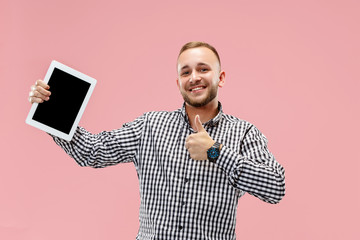 Studio picture of positive man isolated on pink background standing in casual clothes holding tablet and showing it blank screen with happy smile. He advising product or service.