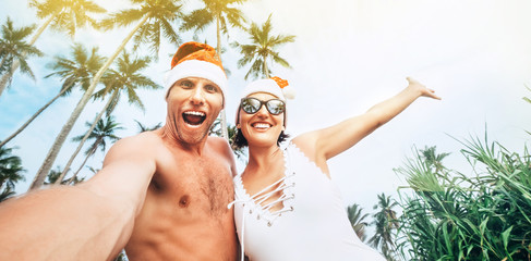 Wall Mural - Christmas image of Cheerful young people couple dressed red Santa hats making selfie with tropic palm trees background .