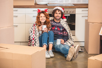 happy young couple holding cups and smiling at camera while sitting between cardboard boxes at chris