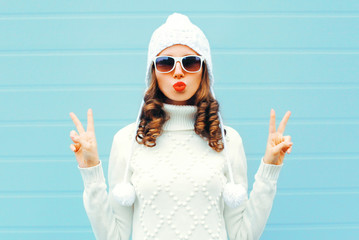 Portrait beautiful woman in white knitted sweater, hat, sends air kiss on blue background