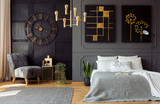 Fototapeta  - Real photo of dark grey bedroom interior with molding and paintings on walls, double bed with pillows, gold lamp and floral armchair
