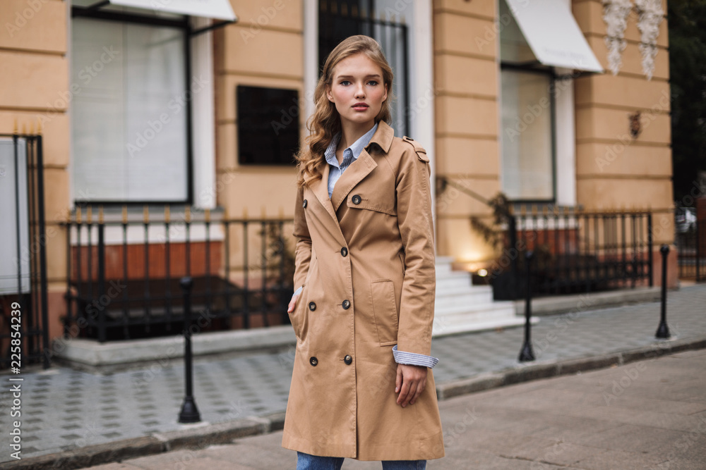 Young attractive woman in trench coat thoughtfully looking in camera while walking around cozy city street - obrazy, fototapety, plakaty 