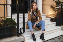 Beautiful Girl In Trench Coat And Jeans Dreamily Looking In Camera Sitting On Little Stairs While Spending Time On Cozy City Street