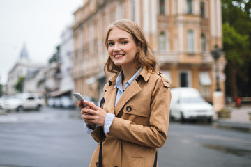 Young cheerful woman in trench coat happily looking in camera holding cellphone in hands while spending time on cozy city street