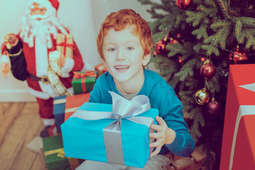 Wall Mural - For you. Handsome boy keeping smile on his face and holding gift box while looking forward