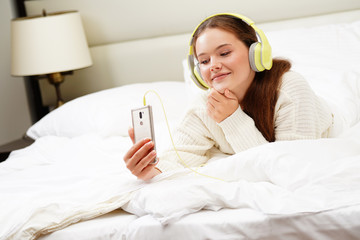 Wall Mural - beautiful attractive young caucasian woman brunette morning in white bed with phone looking in smartphone face smiling dreaming listening music in headphones