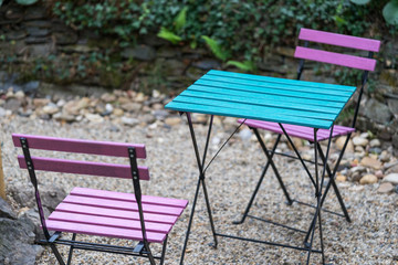 Two pink wooden folding chairs and a square blue table are standing on gravel in the backyard of a cafe in natural light in the evening