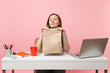 © ViDi Studio - Woman hold brown clear empty blank craft paper bag, sniffing smell work at office with pc laptop isolated on pink background. Food products delivery courier service from shop or restaurant to office.