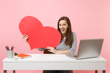 Wall Mural - Young passionate woman holding red empty blank heart sit and work at white desk with pc laptop isolated on pastel pink background. Achievement business career concept. Copy space for advertisement.