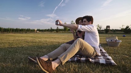 Poster - Young happy loving couple sitting outdoors in the field drinking wine talking with each other take a selfie with mobile phone