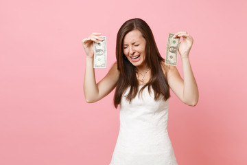 Portrait of upset sad bride woman in white wedding dress crying and holding one dollar bills isolated on pastel pink background. Lack of money. Organization of wedding celebration concept. Copy space.