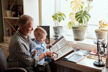 Senior Man And Little Boy Holding And Looking At Family Photo Album In Living Room