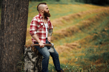 Wall Mural - brutal lumberjack in red shirt with beard holding axe sitting in Forest after work