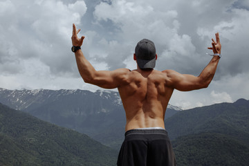 Rear view of muscular bodybuilder with naked tanned torso, arms up outstretched looking at inspirational landscape on mountain top. motivation and inspiration concept