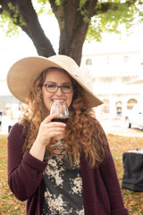 Beautiful young redhead woman, with curly hair, in glasses and a hat, drinking wine at a festival