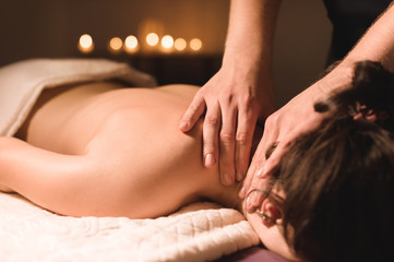 Wall Mural - Men's hands make a therapeutic neck massage for a girl lying on a massage couch in a massage spa with dark lighting. Close-up. Dark Key