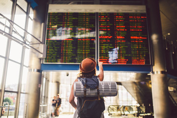 Theme travel public transport. young woman standing back in dress and hat behind backpack camping equipment for sleeping, insulating mat looks schedule on scoreboard airport station hand to the front