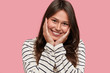 © WHstudio Leushin N - Close up shot of pleasant looking freckled girl smiles positively at camera, keeps hands on cheeks, dressed in striped casual sweater, pleased to recieve compliment, isolated over pink background
