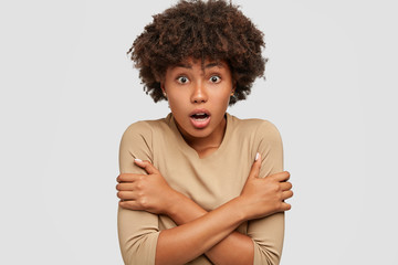 Wall Mural - Omg, I am so scared. Beautiful stunned ethnic woman keeps hands crossed over chest, trembles from fear, tries to defense herself, dressed in casual beige sweater, isolated on white background