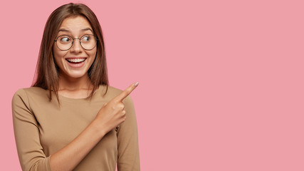 Wall Mural - Studio shot of good looking satisfied brunette woman with toothy broad smile, wears round spectacles, points with index finger aside, shows copy space for your advertising content, being in good mood