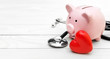 © igorkol_ter - Piggy bank with stethoscope and red heart on white wooden table. Space for text.
