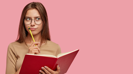 Pensive female writer ponders on new ideas, holds red notebook and pencil near mouth, has thoughtful expression, wears big round glasses, isolated over pink background with copy space on right