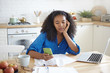 © shurkin_son - Beautiful young Afro American female holding cell phone using online banking application while paying domestic bills. Pretty girl planning family budget sitting at table with papers and gadgets