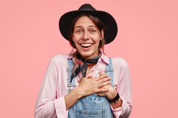 Moved beautiful smiling gardener keeps both hands on heart, being pleased by positive compliment, dressed in stylish clothes, poses over pink background, thankful for warm words, wears black hat