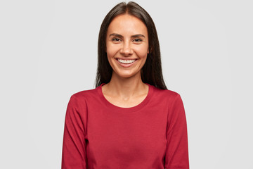 Photo of satisfied brunette woman with dark hair, has pleasant smile, being in good mood, wears red casual sweater, isolated over white background. Tanned lady rejoices positive moment in life