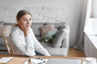 © shurkin_son - Elderly sixty year old female teacher sitting at her workplace with laptop, coffee, glasses and textbooks on desk, having break between online lessons, looking at camera with serious expression