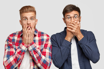 Wall Mural - Stunned shocked two guys gasp from fear, cover mouthes with both hands, being astonished with terrible event, stand next to each other, have stylish haircuts, isolated on white blank studio wall