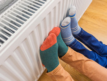 Woman And Child Wearing Colorful Pair Of Woolly Socks Warming Cold Feet In Front Of Heating Radiator In Winter Time. Electric Or Gas Heater At Home.