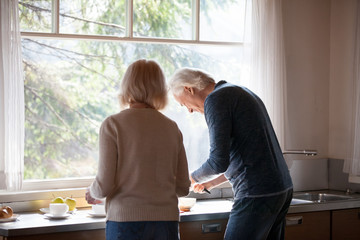 Rear view at middle aged loving couple preparing breakfast together in the kitchen standing at big window, caring mature husband helping senior wife to cook morning meal, old people at home lifestyle