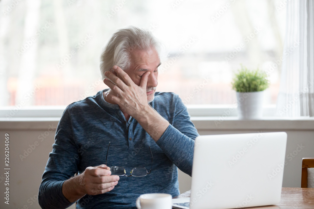 Fatigued mature old man taking off glasses suffering from tired dry irritated eyes after long computer use, senior middle aged male feels eye strain problem or blurry vision working on laptop at home - obrazy, fototapety, plakaty 