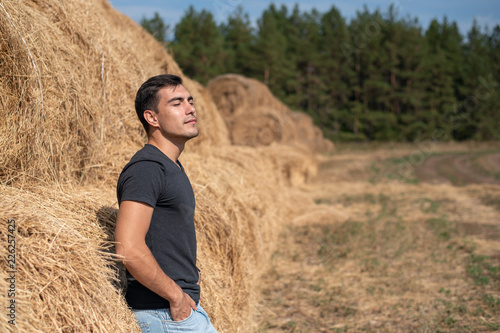 Fotografie, Obraz A young man in a gray t-shirt and jeans stands at a haystack, breathing clean ai