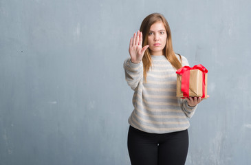 Sticker - Young adult woman over grey grunge wall holding a present with open hand doing stop sign with serious and confident expression, defense gesture