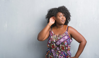 Canvas Print - Young african american woman over grey grunge wall wearing colorful dress smiling with hand over ear listening an hearing to rumor or gossip. Deafness concept.