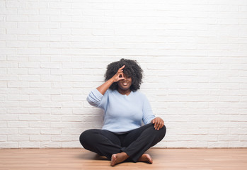 Canvas Print - Young african american woman sitting on the floor at home doing ok gesture with hand smiling, eye looking through fingers with happy face.