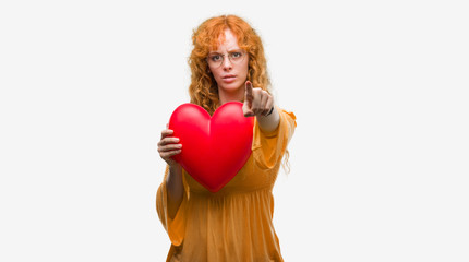 Poster - Young redhead woman in love holding red heart pointing with finger to the camera and to you, hand sign, positive and confident gesture from the front