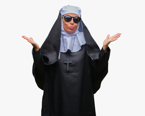 Middle age senior catholic nun woman wearing sunglasses over isolated background clueless and confused expression with arms and hands raised. Doubt concept.