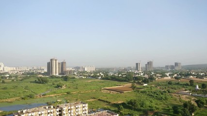 Wall Mural - Aerial panning shot o fgurgaon city showing the old farmlands and skyscraper buildings in the distance. The grassy lands, farms, lakes contrast sharply with the sky scrapers multistory buildings in