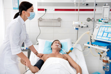 Young female doctor in protective face mask touching hand and shoulder of patient while looking at monitor of breathing machine. Man lying in hospital bed with endotracheal tube in the mouth